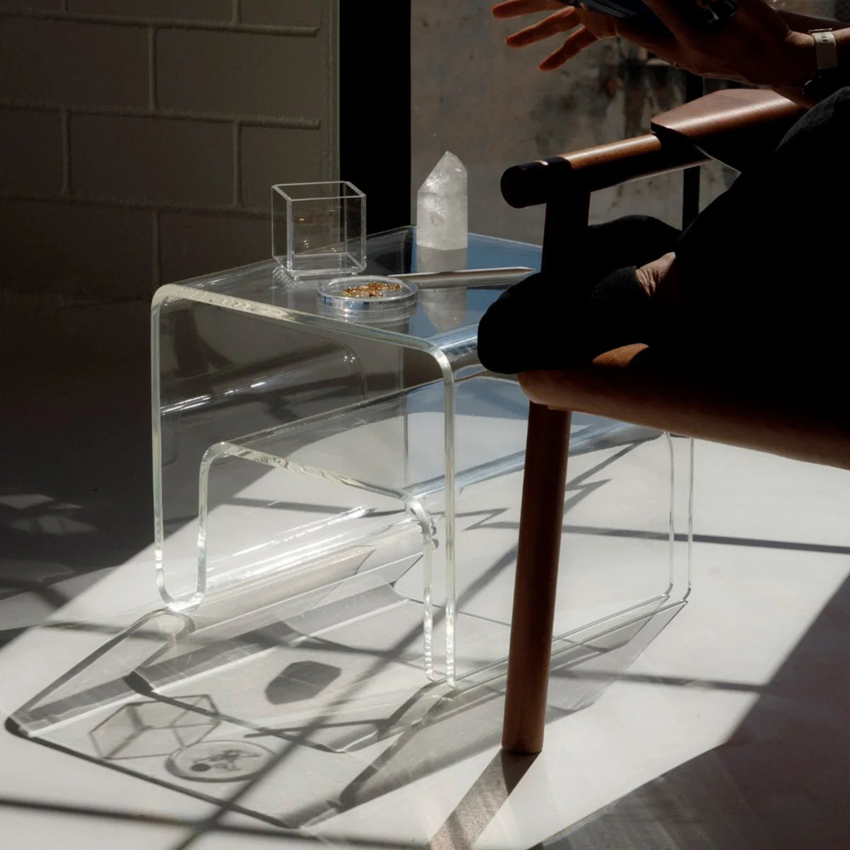 Mesa lateral Vértis em acrílico cristal, ao lado de pessoa sentada, com copo e jarra.