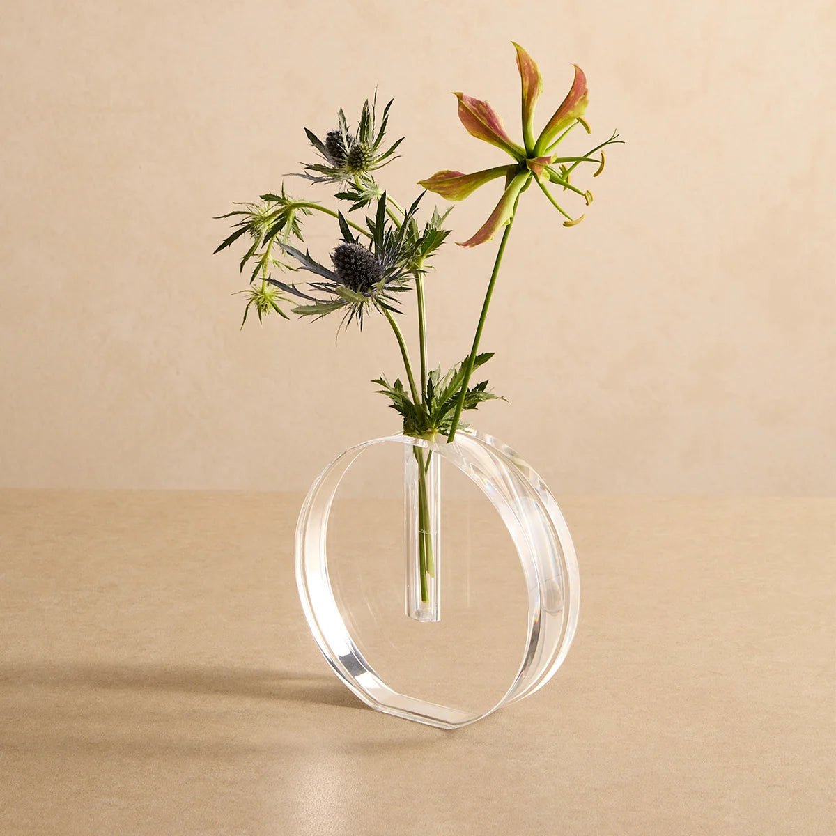 Vaso Sinué em acrílico cristal, com flores sobre fundo bege.
