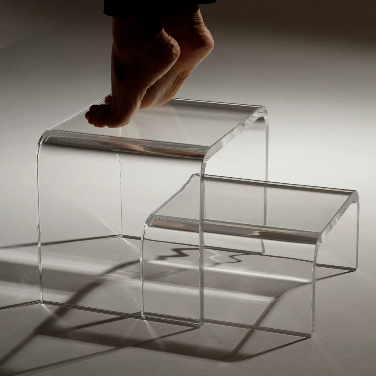 Escada modular Altea em acrílico cristal, com pé descalço sobre o degrau superior.
