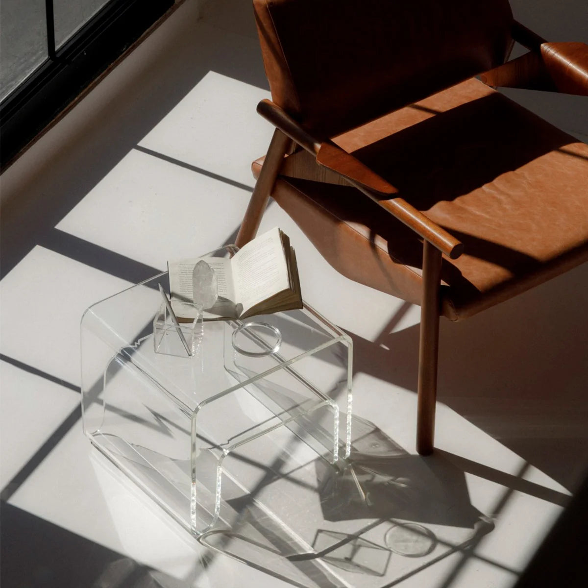 Mesa lateral Vértis em acrílico cristal, vista de cima com livro ao lado de poltrona.